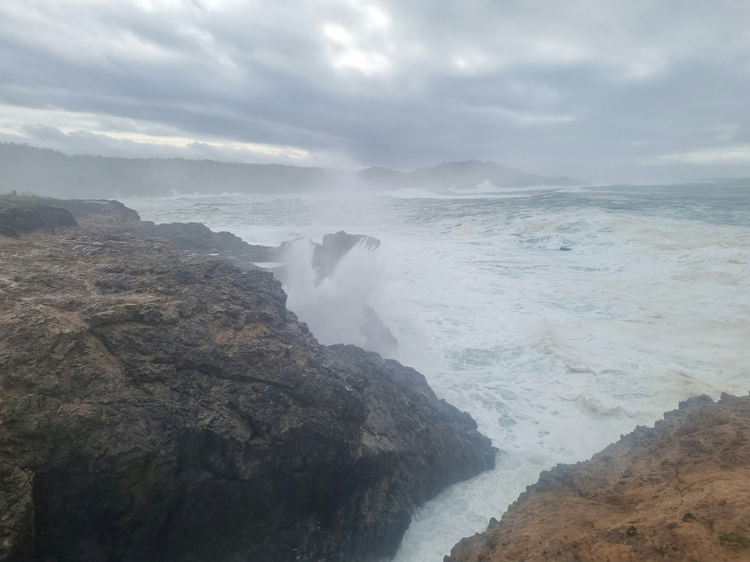 Surf breaking white over a dark rocky headland on the Northern California coast under heavy weather