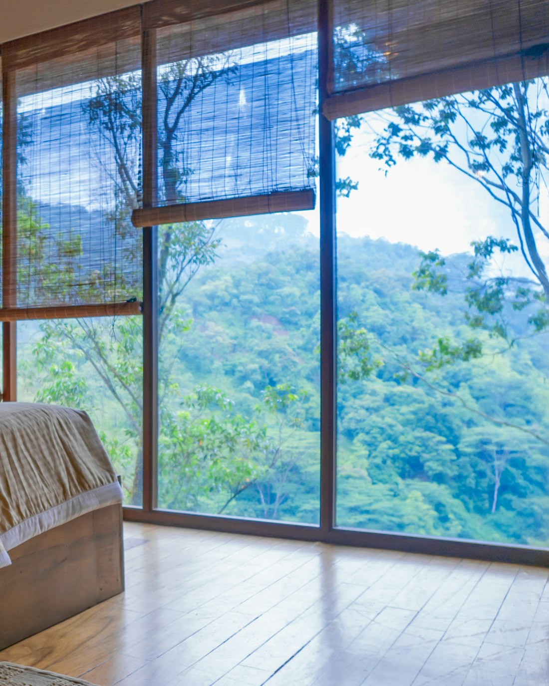 A bed made up in pale linen beside a window wall looking into the forest